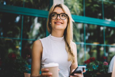 Sıcak içecek ve modern akıllı telefon fincan ile Pretty genç kadın gülümseyen ve açık kafede serbest iş yaparken uzak bakıyor