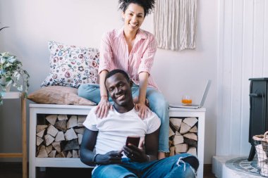 Neşeli genç etnik çift birlikte gülümseyip mutlu anların tadını çıkarırken şöminenin yanında kırsal bir oturma odasında oturup akıllı telefon kullanıyorlar.