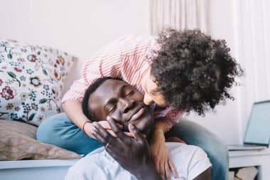 Çizgili tişörtlü, etnik kıvırcık saçlı kadın, oturma odasında şömineli otururken arkadan sarılıp yanaktan öpüyor.