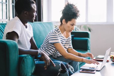 Siyahi adamın odaklanmış kız arkadaşına dizüstü bilgisayar kullanarak bakarken evdeki pencereye karşı rahat bir koltukta oturması.