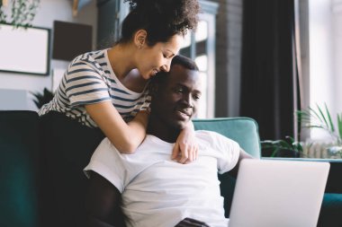İyi ki Afro-Amerikalı bir kadın dizüstü bilgisayarla etnik erkeği kucaklıyor ve rahat koltuğun arkasında durup evinde dinlenirken gülümsüyor.