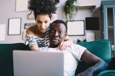 Hayretler içinde kalan Afrikalı Amerikalı erkek ve kadın modern dairenin rahat odasında dinlenirken dizüstü bilgisayardan şok edici videolar izliyorlar.