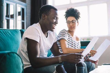 Neşeli Afrikalı Amerikalı adam, rahat oturma odasındaki kanepede ciddi kız arkadaşıyla serbest çalışırken kâğıdı inceliyor ve gülüyor.