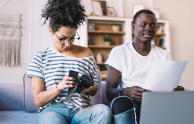 Düşünceli Afrikalı Amerikalı bir adam kanepeye oturup gazete okurken, etnik bir kadın online ödeme için kredi kartı kullanıyor.