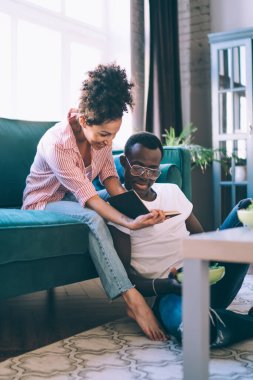 Yetişkin, etnik çeşitlilikte bir çift yemek ve içecekle masaya otururken gülüyor ve modern apartmanda fotoğraf albümü yapmayı düşünüyor.