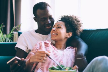 Çok ırklı bir çift rahat koltukta rahat rahat oturup güneşli bir günde gülümseyen ve cep telefonlarını karıştıran sağlıklı kahvaltının tadını çıkarıyorlar.