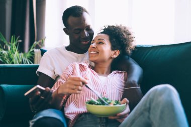 Çok ırklı bir çift rahat koltukta rahat rahat oturup güneşli bir günde gülümseyen ve cep telefonlarını karıştıran sağlıklı kahvaltının tadını çıkarıyorlar.