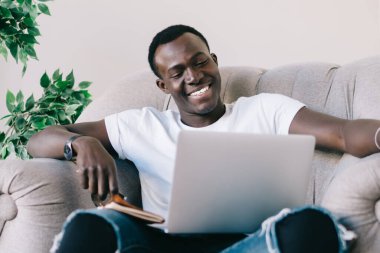 Genç, yırtık kot pantolonlu, kitap seyreden ve modern bir dairede gülümseyen Afro-Amerikan bir adam.