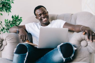 Genç, yırtık kot pantolonlu, kitap seyreden ve modern bir dairede gülümseyen Afro-Amerikan bir adam.