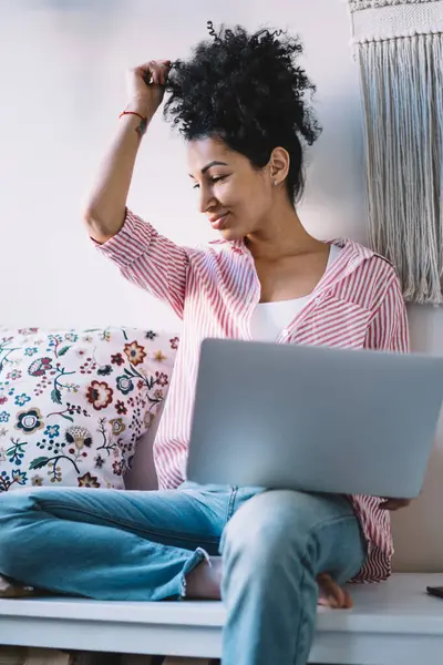 Modern odada dizüstü bilgisayar kullanırken ve başka tarafa bakarken, rahat ve rahat giyinen, rahat bir etnik kadın.