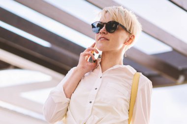 Kampüste boş sırasında modern hücresel Gadget üzerinden uluslararası konuşma yapmak için Roaming 4g internet kullanarak şık güneş gözlüğü büyüleyici hipster kız, kadın müşteri hizmetine çağrıda