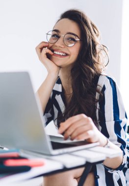 Çekici, soyulmuş takım elbiseli, gözlüklü, dizüstü bilgisayarın önünde oturan, çenesine yaslanıp gülümseyen neşeli genç bir kadın.