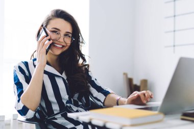 Koyu renk saçlı, neşeli, dişlek gülüşlü, cep telefonuyla konuşan ve modern aydınlık odada bilgisayarla çalışan güzel bir kadın.