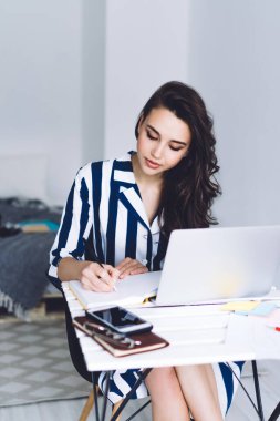 Oldukça sakin, düşünceli bir kadın dizüstü bilgisayarlı telefonla ve kırtasiyeyle masada otururken not defterine not alıyor. Işıklı yatak odasında beyaz duvarlı, bulanık arka planda.