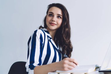 Güzel gülümseyen, düşünceli bir bayanın elinde kalemle dizüstü bilgisayarla masaya otururken yüzünü çevirmesi ve gündüz vakti beyaz duvara karşı portakal suyu ve siyah kamışla cam camlara vurması.