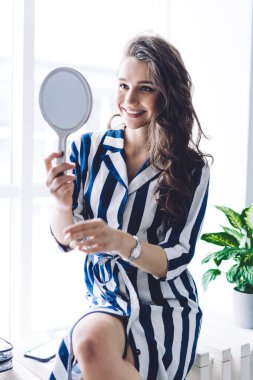 Moda kıyafetleri içinde çekici bir kadın pencere pervazında oturuyor ve evde ayna ve kozmetik ürünleriyle tuvalet yapıyor.