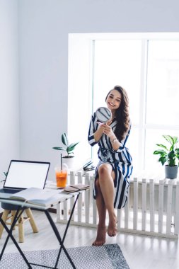 Çekici, genç, neşeli, çizgili elbiseli, el aynasına bakan, pencerede bacak bacak bacak üstüne atmış bir kadın.