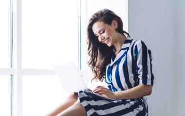 Beyaz modern odada pencerenin yanında oturan ve bilgisayarda yazan çizgili elbiseli neşeli, mutlu bir kadın.