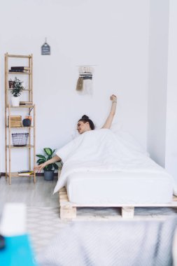 Genç bir kadın, minimalist beyaz bir odada tek başına uyanır ve yatağın yanındaki rafta bir şeye uzanmaya çalışırken ellerini uzatır.