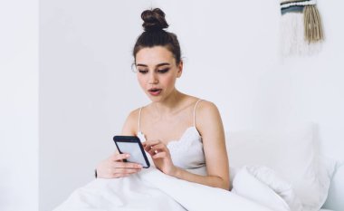Odaklanmış bir kadın, beyaz battaniye ve yastık ile rahat yumuşak yatak üzerinde uyanır ve ışık odasında minimalist iç mekan ile akıllı telefon sörfü yapar.