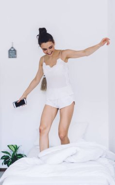 Gece elbiseli mutlu bir kadın, rahat bir yatakta uzanmış, ellerini uzatmış, akıllı telefon kullanıyor ve modern iç mekanı seyrediyor.