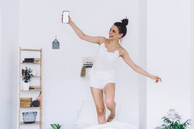 Beyaz pijamalı neşeli genç bir kadın boş ekranlı akıllı telefon taşıyor ve modern yatak odasında beyaz duvarlı yatağa atlıyor.