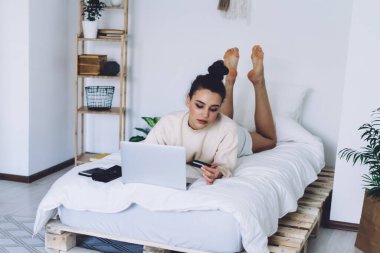 Ev kıyafetleri içinde çekici bir esmer, saçı topuz, kredi kartına bakıyor ve modern odada el yapımı bir yatakta dinlenirken dizüstü bilgisayar kullanarak internetten alışveriş yapıyor.