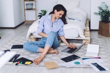 Gündelik giysiler içinde, aydınlık yatak odasında, fosforlu kalemler ve çizelgeler arasında dizüstü bilgisayar kullanarak yerde oturan bir kadın.