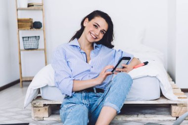 Gündelik kıyafetlerle gülümseyen, notlar alan, akıllı telefonları ve laptopları karıştıran ve modern apartmanda çıplak ayakla yerde oturan kendine güvenen bir kadın.