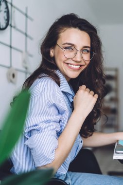 Gözlüklü, mavi gömlekli, beyaz ekranlı, dizüstü bilgisayarını karıştıran ve kameraya bakan güzel bir kadının yan görüntüsü.