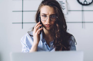 Buruşuk burun köprüsü ve gözlüklü endişeli kadın işçiler. Birden fazla iş üzerine yoğunlaşarak akıllı telefondan konuşuyor ve ofiste dizüstü bilgisayar kullanıyor.