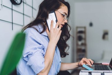 Gözlüklü, sıradan giyinmiş, dizüstü bilgisayar kullanan ve ev telefonuyla konuşan genç bir kadının yan görüntüsü.