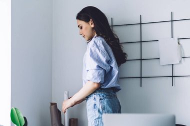 Mavi kot pantolon ve bluz giymiş, ofis odasının köşesinde duvardan duvara duran ve beyaz kağıt rulo alan kendine güvenen genç kadının yan görüntüsü.