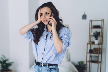 Olaylı, kasvetli, siyah saçlı, rahat kot ve tişörtlü bir kadın. Cep telefonuyla konuşuyor, elleri başının üstünde güneşli bir yatak odasında ve başka tarafa bakıyor.