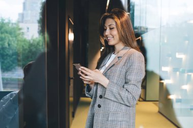 Modern ofis binasının koridorunda dururken akıllı telefondan mesaj atan mutlu etnik kadın girişimcinin cam tarafından bakıldığında,