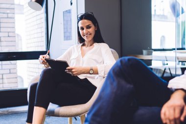 Mutlu ve neşeli bir kadın meslektaşının yanında oturuyor ve toplantı sırasında notlar alıp kameraya bakarken gülümsüyor.