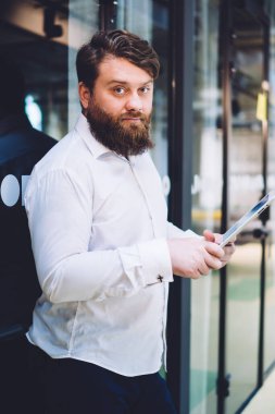 Ciddi sakallı bir yönetici, modern ofis binasında cam kapıya yaslanırken kameraya bakıyor ve tablet üzerinde çalışıyor.