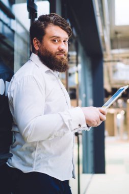 Modern ofis binasında cam kapıya yaslanırken kameraya bakan ve tablet üzerinde çalışan ciddi sakallı erkek yöneticinin yan görüntüsü bulanık arka planda.