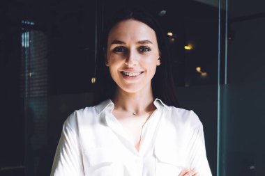 Parlak odada kollarını kavuşturup kameraya bakarken gülümseyen resmi giyinmiş neşeli ve mutlu bir bayan.