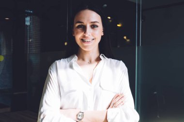 Parlak odada kollarını kavuşturup kameraya bakarken gülümseyen resmi giyinmiş neşeli ve mutlu bir bayan.