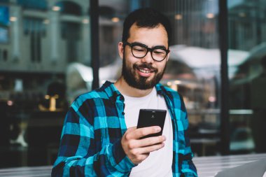 Genç serbest çalışan akıllı telefondaki uyarıyı uzaktan kontrol eden kablosuz ücretsiz internet kullanan hipster adam dizüstü bilgisayarda oturup sosyal ağlarda sohbet ediyor.