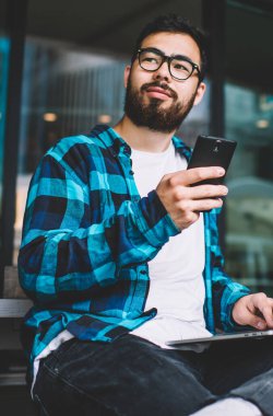 Kazak genç adam elinde akıllı telefon aygıtı tutarken modern dizüstü bilgisayarda kablosuz internet bağlantısı kullanıyor, erkek blogcu netbook cihazının başında oturan cep telefonu uygulamasını güncelliyor