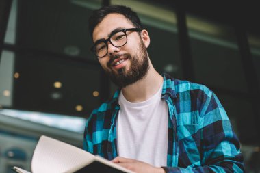 Optik gözlüklü neşeli kazak adamın portresi kameraya gülümseyen göz korumasını sağlamak için eğitim kitapçığında ders programını planlarken, pozitif erkek öğrenci not defteri tutuyor.