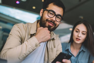 Gözlüklü neşeli sakallı hippi adamın portresi kameraya gülümsüyor ve güzel beyaz kız arkadaşıyla vakit geçirmekten zevk alıyor yanında oturuyor ve cep telefonunun ekranına bakıyor.