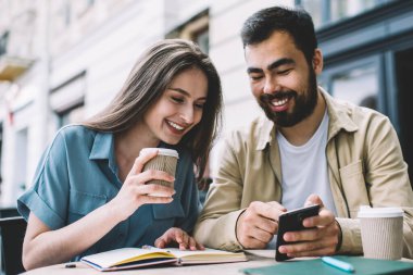 Neşeli erkek ve bayan meslektaşlar sosyal ağlardan komik yayınlar okuyor ve kahve molası sırasında sokak kafeteryasında sınav hazırlığı sırasında gülüyor, mutlu arkadaşlar içeriği tartışıyor.