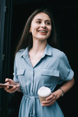 Neşeli kadın blogcu elinde kafein içeceğiyle kafeteryadan çıkarken keyfine bakıyor ve gülümsüyor, gündelik giyinmiş, elinde kağıt bardak ve cep telefonu tutan neşeli bir kadın.