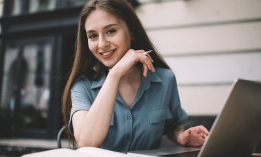 İlginç olumlu makaleler yaratmak için dizüstü bilgisayar ve ders kitabı kullanan çekici gülümseyen kadın metin yazarının portresi, Kafkasyalı serbest yazarların sokak kafeteryasında zaman geçirmesi
