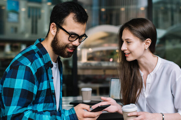 Couple having discussion while making online booking on smartphone browsing webpage, male and female best friends communicating while searching media in social networks resting together in coffee shop