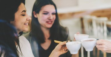 Gündelik modaya uygun giyinmiş, birlikte vakit geçirmekten keyif alan, kahve fincanlarıyla tezahürat yapan, yeni başlayanlar projesinde işbirliği yapan, iç mekanda sosyalleşen mutlu bayan meslektaşlar.