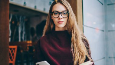 Optik gözlüklü ciddi bir kadın okuyucunun portresi eğitim için göz koruması, not defteri ve akıllı telefon cihazıyla elinde kahve dükkanında oturan genç bir kadın.